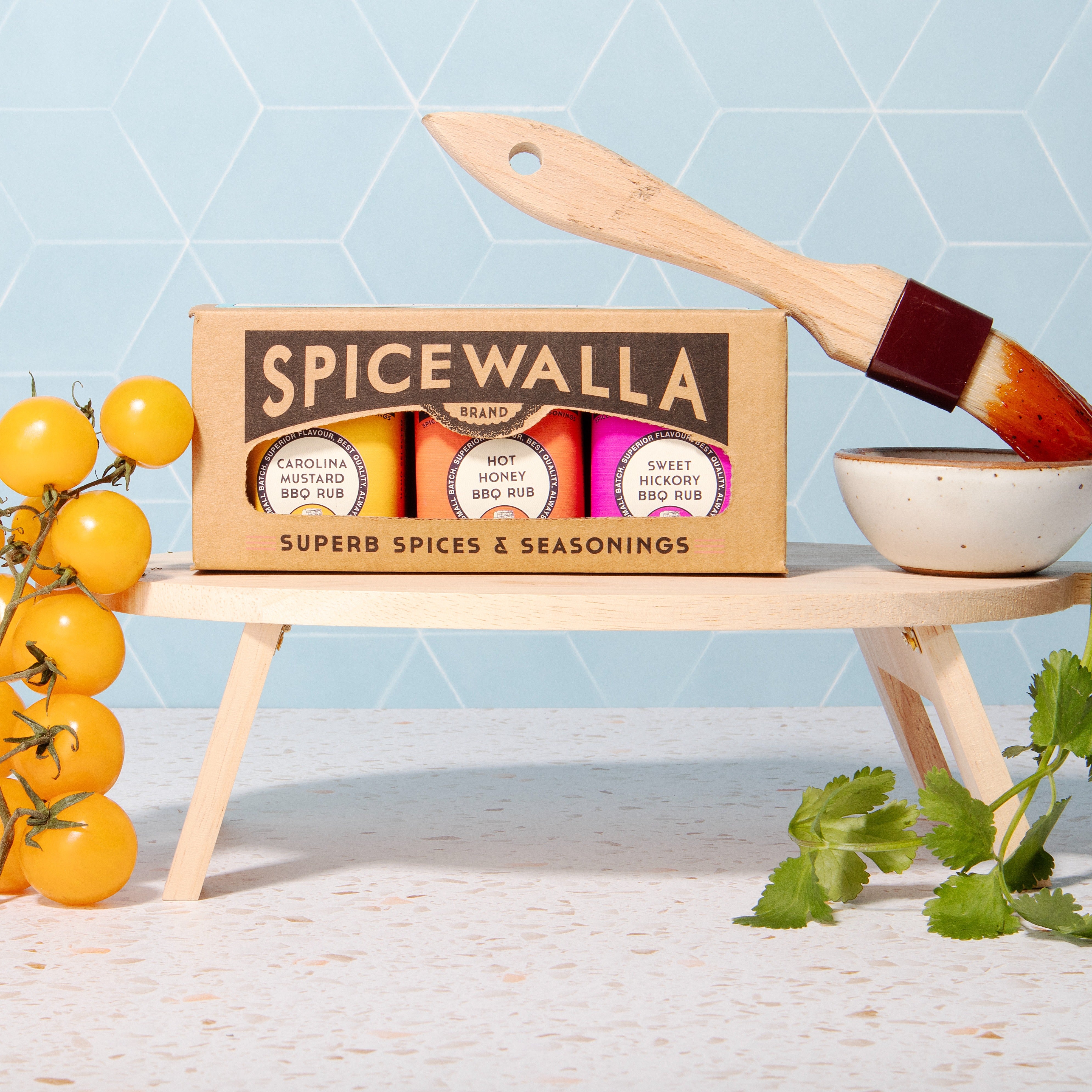 Spicewalla spice set with a wooden shelf, yellow tomatoes, and a brush on a light blue geometric patterned background.