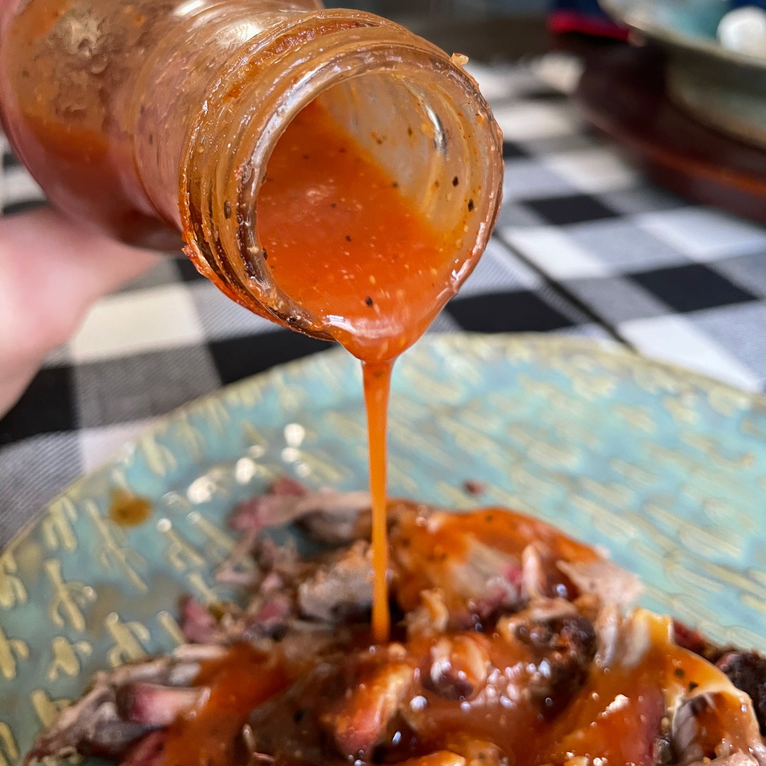 Hot sauce being poured from a jar onto a dish on a checkered tablecloth.