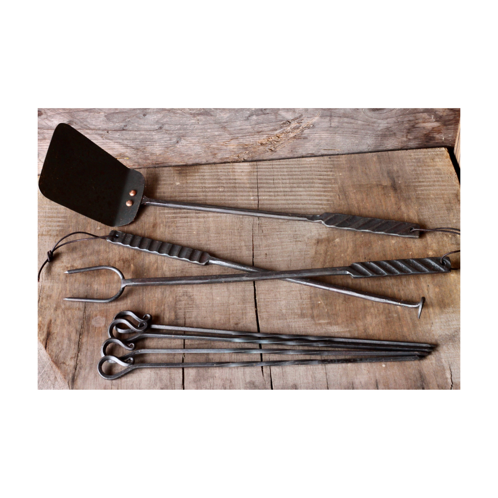 Set of barbecue tools including a spatula, tongs, and skewers on a wooden surface.