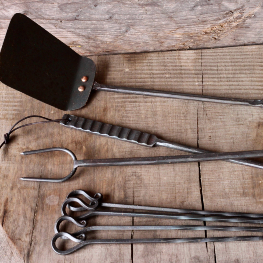 Set of barbecue tools including a spatula, tongs, and skewers on a wooden surface.