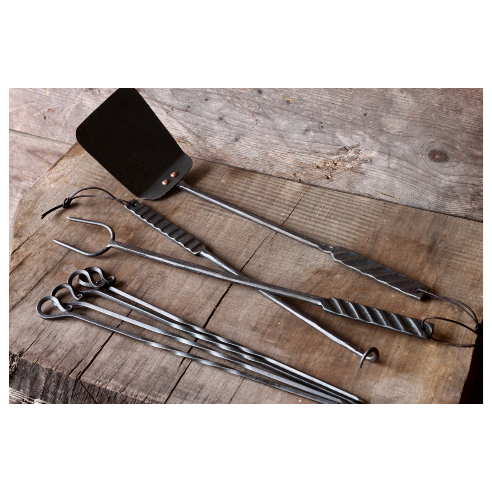 Set of barbecue tools including a spatula, tongs, and skewers on a wooden surface.