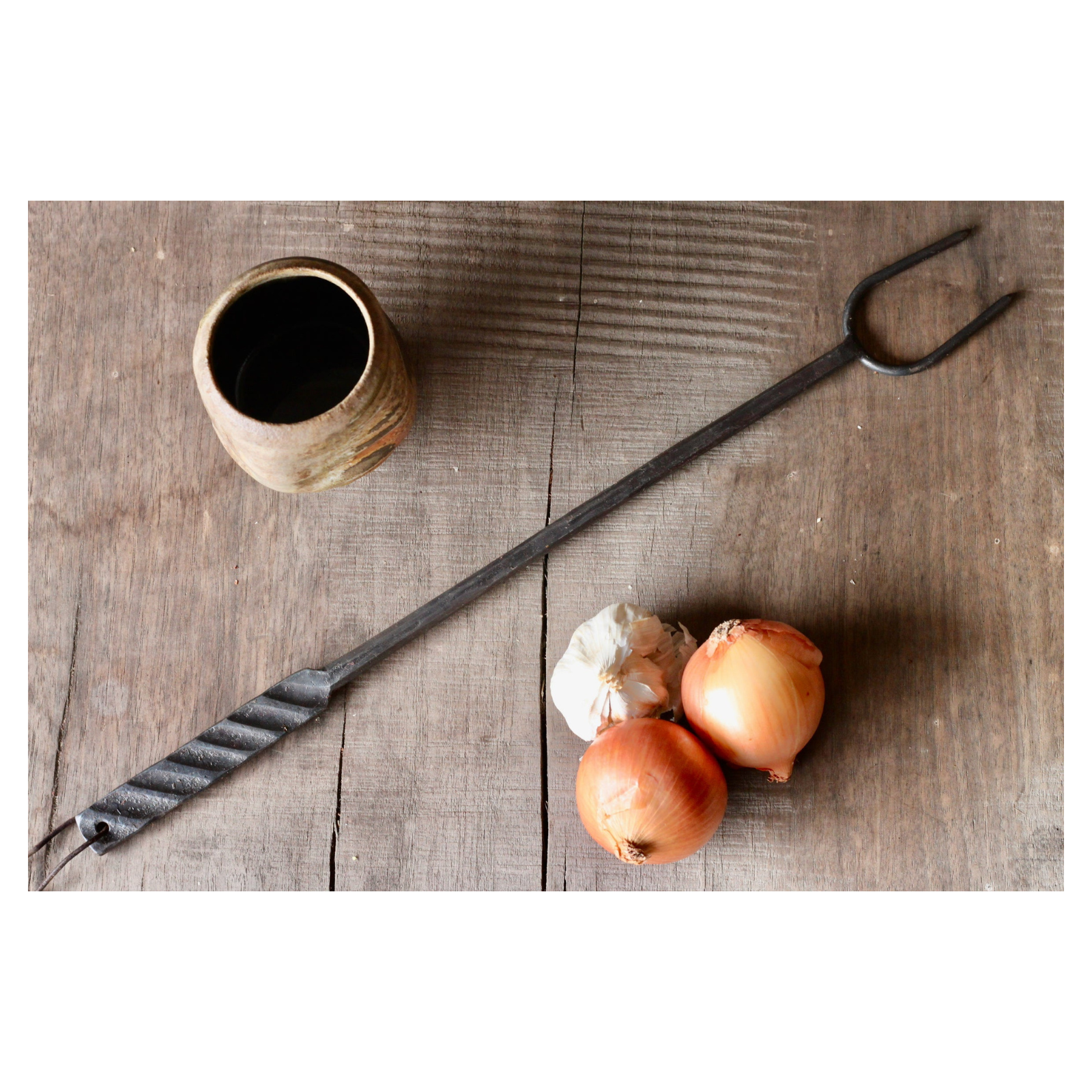Long-handled metal tool, onions, and a ceramic container on a wooden surface