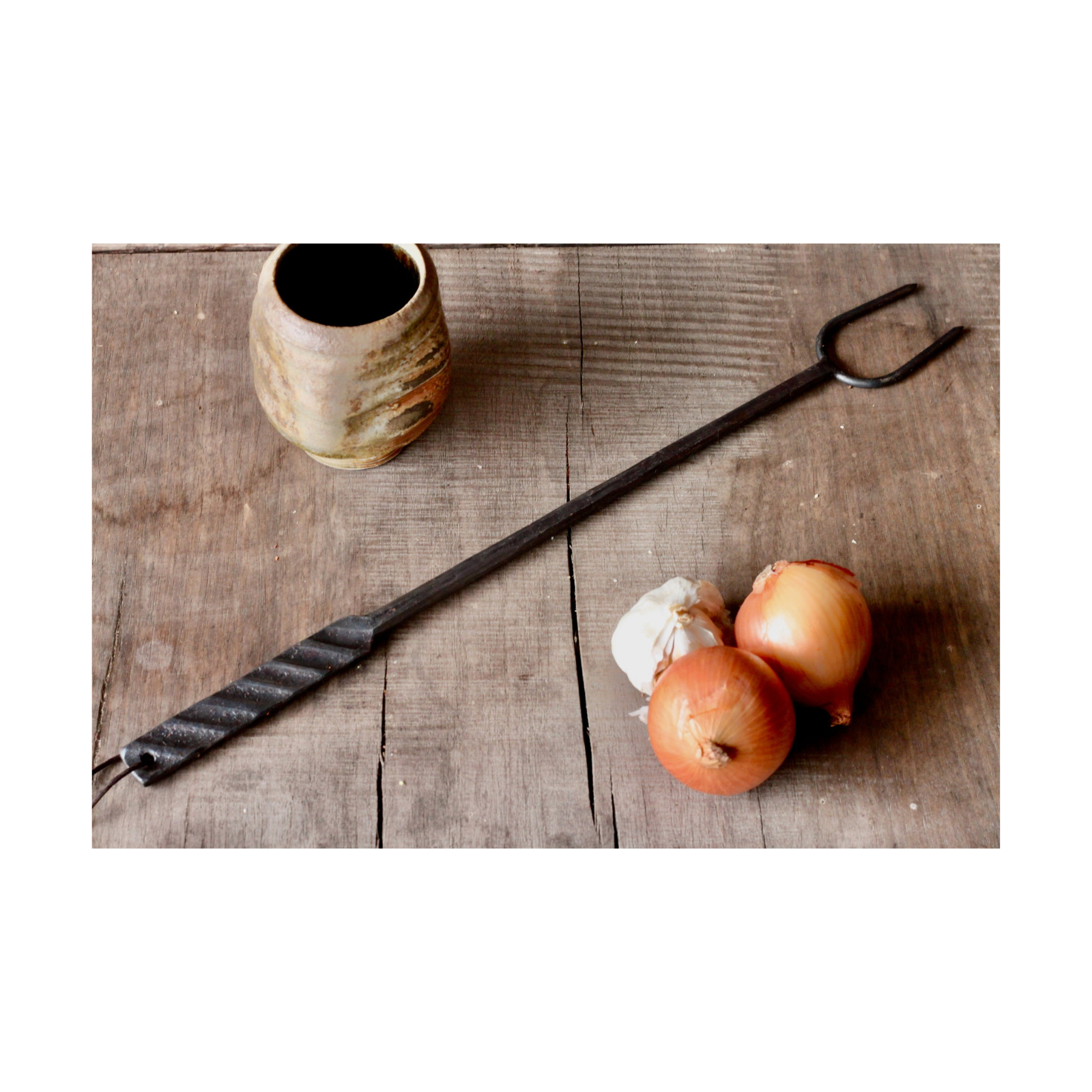 Black metal tool, possibly a fireplace poker, on a wooden surface with onions and a ceramic container.