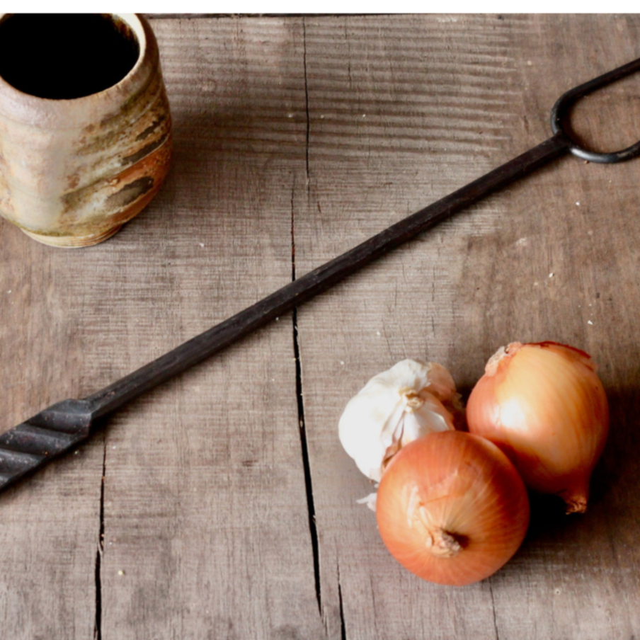 Black metal tool, possibly a fireplace poker, on a wooden surface with onions and a ceramic container.